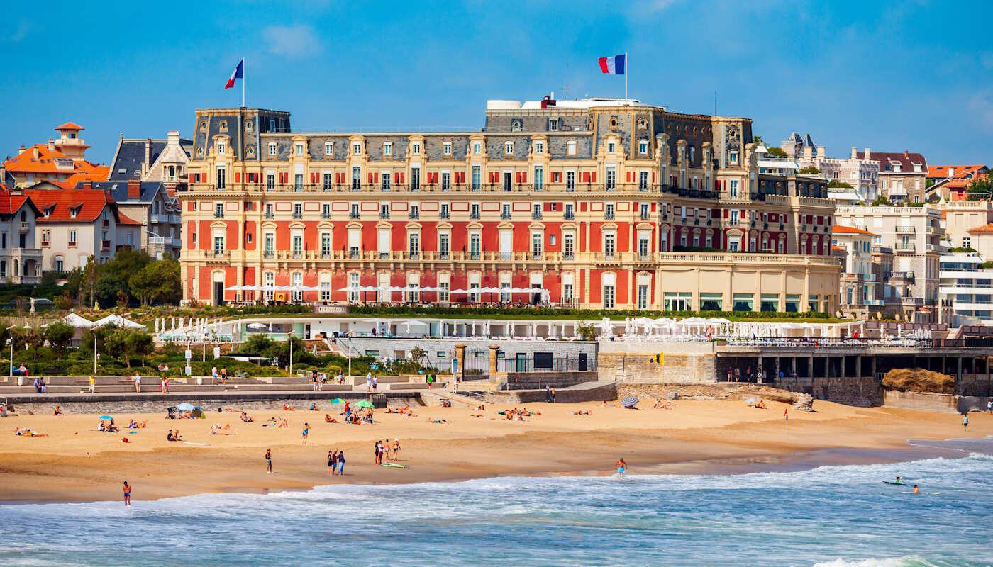 Hotel du Palais in Biarritz, Frankreich Das Bild zeigt die Grande Plage in Biarritz mit dem imposanten roten Hôtel du Palais direkt an der Atlantikküste. Vor dem historischen Luxushotel erstrecken sich ein breiter Sandstrand, die charakteristischen Atlantikwellen und die elegante Küstenarchitektur des berühmten Badeortes. Der Himmel ist klar und blau.