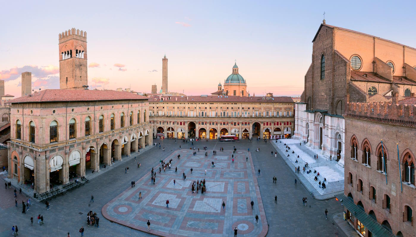 Piazza Maggiore in Bologna, Italien Das Bild zeigt die beeindruckende Piazza Maggiore im historischen Zentrum von Bologna, einem der bekanntesten Plätze der Stadt. Zu sehen sind die markante Basilica di San Petronio auf der rechten Seite, der Palazzo dei Banchi im Hintergrund sowie mehrere mittelalterliche Türme, während Menschen über den weitläufigen Platz schlendern.
