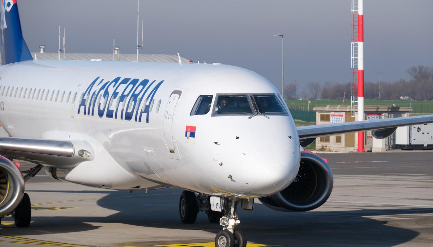 Air Serbia Maschine Ein Flugzeug der Airline Air Serbia steht auf dem Rollfeld eines Flughafens. Die Maschine ist weiß und trägt den Schriftzug „Air Serbia“ auf dem Rumpf. Im Hintergrund sind Startbahn, Gebäude und ein rot-weißer Mast zu sehen.