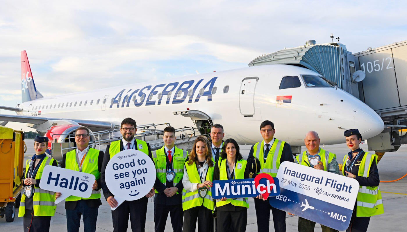 Erstflug AirSERBIA Ein Flugzeug von Air Serbia steht am Gate eines Flughafens. Davor posiert eine Gruppe von Personen in Warnwesten und Uniformen mit Schildern. Auf einem Schild steht „Inaugural Flight 22 May 2026“ und „Munich“.