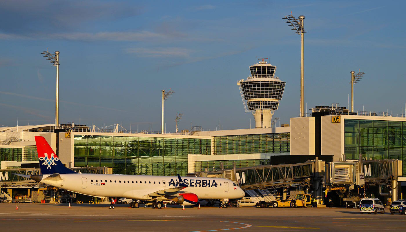 AirSERBIA am Flughafen München Ein Flugzeug von Air Serbia steht am Terminal auf dem Rollfeld. Im Hintergrund sind ein Kontrollturm, Gebäude und Fluggastbrücken zu sehen. Fahrzeuge und Bodenmarkierungen zeigen den aktiven Flughafenbetrieb.