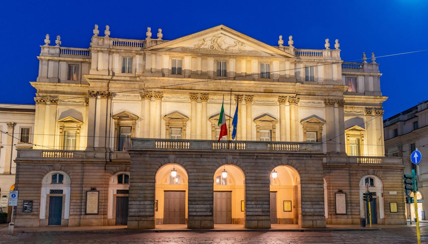 Teatro alla Scala in Mailand, Italien Das Bild zeigt das Teatro alla Scala in Mailand, Italien, ein berühmtes Opernhaus mit klassizistischer Fassade. Zu sehen ist die beleuchtete Front mit Säulen, Balkonen und Flaggen, aufgenommen in den Abendstunden.