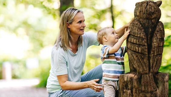 Auf dem Walderlebnispfad im Freisinger Forst kniet eine Frau neben einem Kleinkind. Das Kind streckt die Hand aus und berührt eine große geschnitzte Eulenfigur aus Holz.