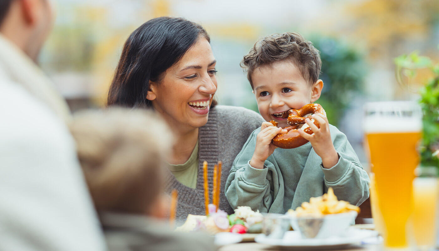 Familienzeit im Airbräu Familienzeit im Airbräu: Kind isst eine Brezel an einem Tisch im Biergarten am Flughafen München, während seine Mutter lächelnd daneben sitzt.