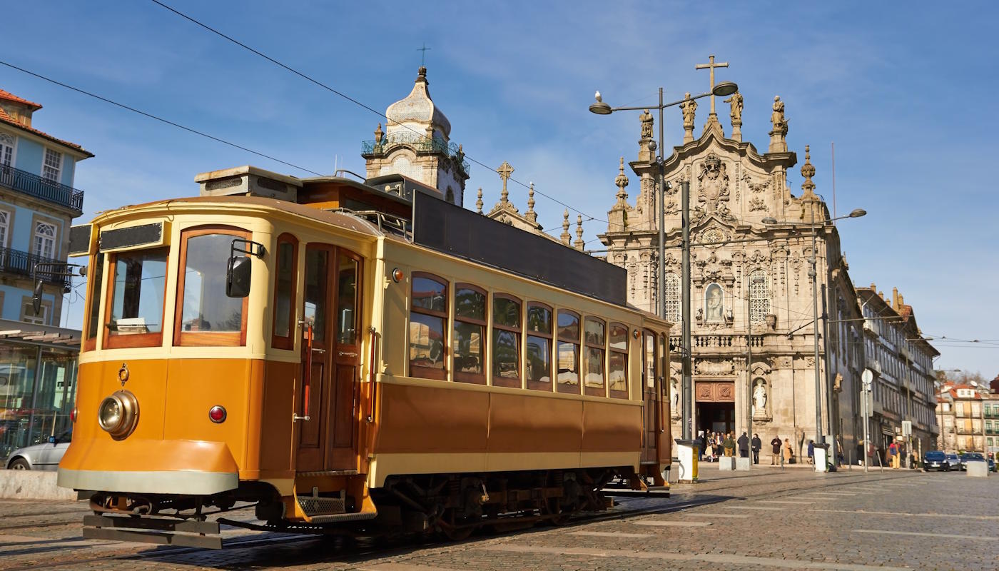 Straßenbahn in Porto, Portugal Das Bild zeigt die historische Altstadt von Porto in Portugal: Im Vordergrund fährt eine gelb-braune, nostalgische Straßenbahn über Kopfsteinpflaster, während im Hintergrund eine reich verzierte Barockkirche mit steinerner Fassade und Türmen zu sehen ist. Die Szene wirkt sonnig und ruhig, mit einigen Passanten auf dem Platz, Oberleitungen über der Straße und historischen Gebäuden, die das traditionelle Stadtbild prägen.