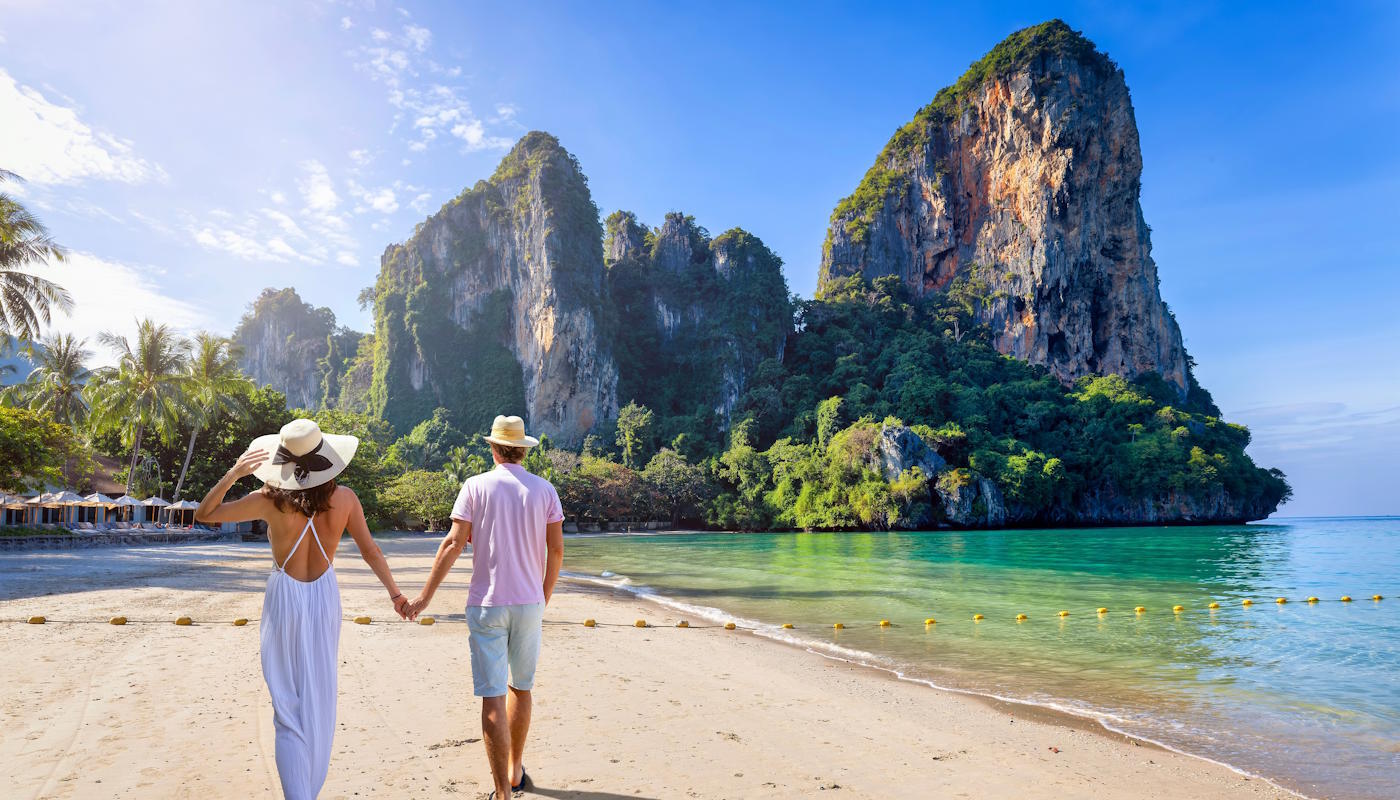 Junges Paar am Strand auf der Insel Krabi in Thailand Ein Paar geht Hand in Hand über einen hellen Sandstrand auf der thailändischen Insel Krabi auf eine türkisfarbene Bucht zu, im Hintergrund ragen steile, dicht bewachsene Kalksteinfelsen aus dem Meer unter einem klaren blauen Himmel empor.