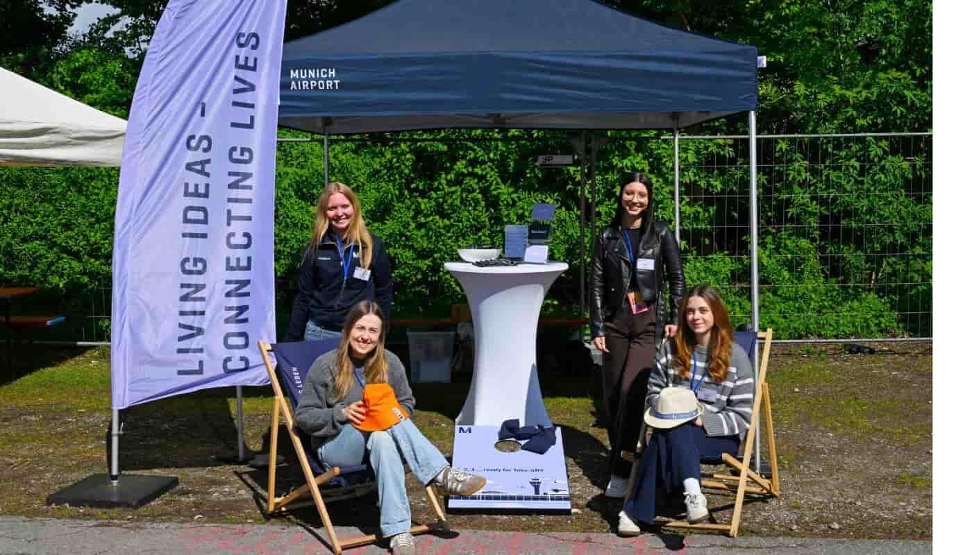 Flughafen München beim FünfSieben Festival – Engagement für Jugendliche und die Region Vier junge Mitarbeiterinnen des Flughafens München begrüßen Besucherinnen und Besucher am Informationsstand des FünfSieben Festivals. Vor dem Pavillon mit der Botschaft „Living Ideas – Connecting Lives“ entsteht eine offene und einladende Atmosphäre für Begegnung, Austausch und gemeinsames Engagement.