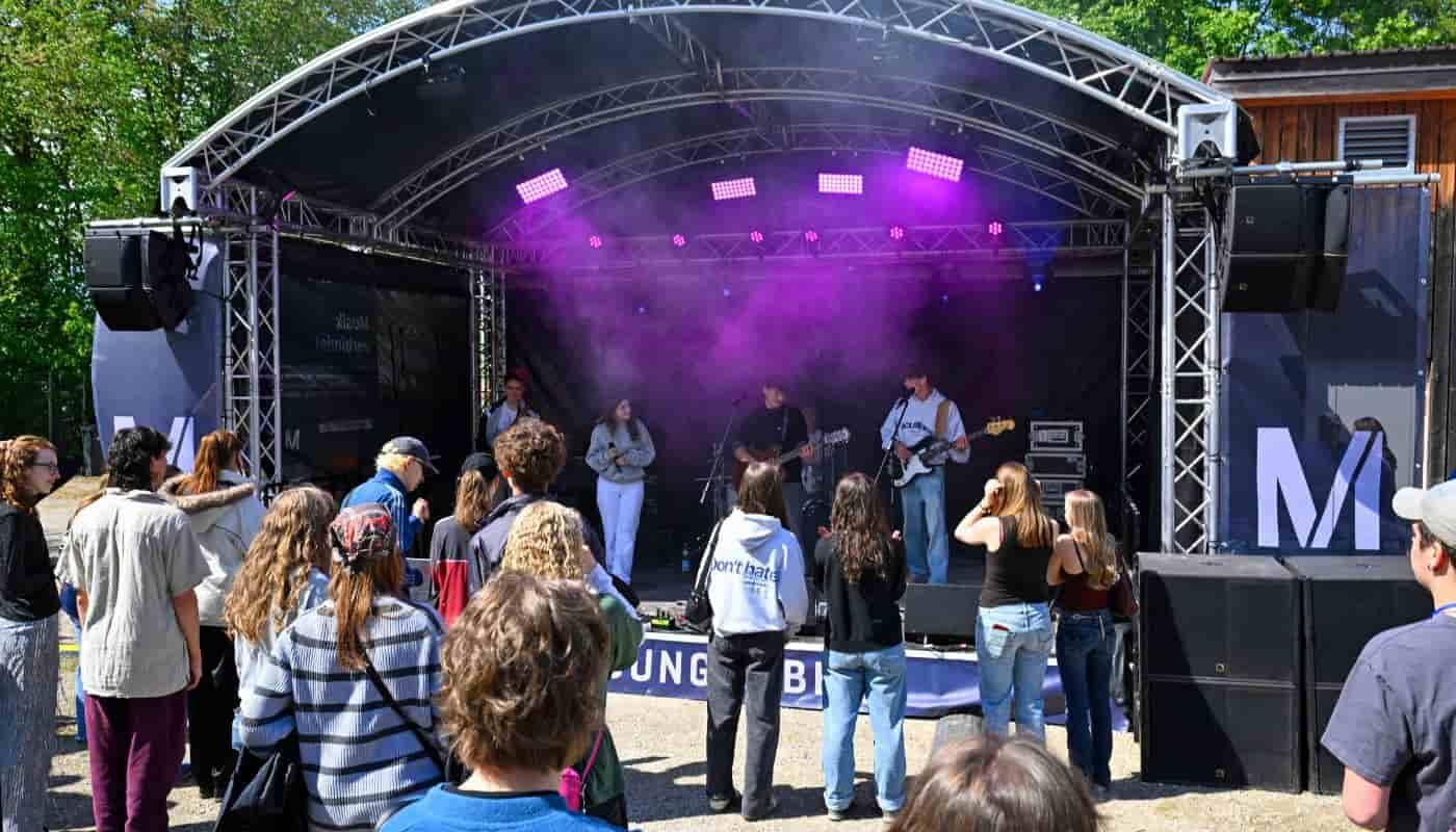 FünfSieben Festival am Karlsfelder See – Flughafen München fördert Jugendkultur Junge Besucherinnen und Besucher erleben die Live-Atmosphäre des FünfSieben Festivals vor einer Open-Air-Bühne am Karlsfelder See, während eine Band auftritt und Musik, Gemeinschaft und Jugendkultur verbindet.