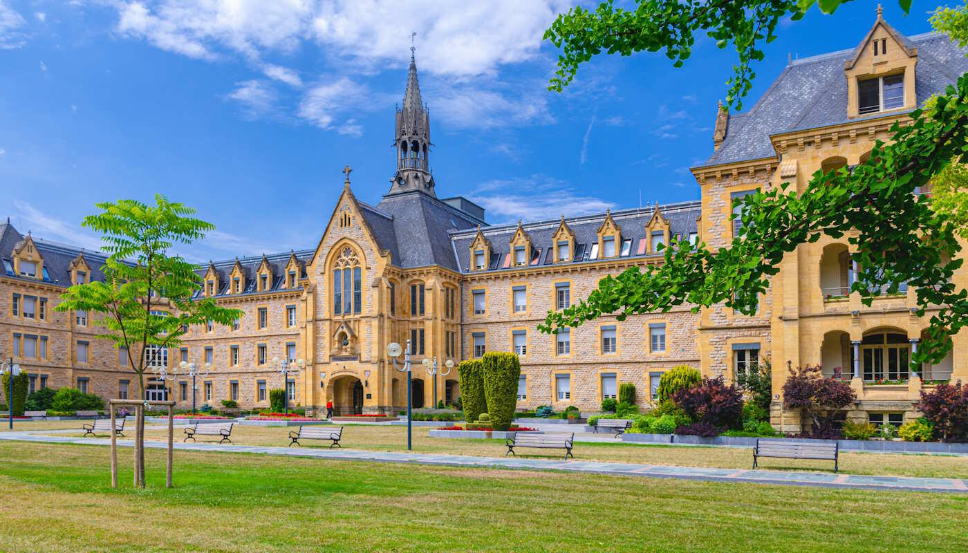 Altstadt von Luxemburg Stadt, Luxemburg In Luxemburg steht ein großes historisches Gebäude aus hellem Stein mit vielen Fenstern, einem spitzen Turm und einem gepflegten Park mit Rasenflächen, Bäumen und Sitzbänken davor. Über dem Gebäude spannt sich ein leuchtend blauer Himmel mit weißen Wolken, wodurch die Szene freundlich und einladend wirkt.