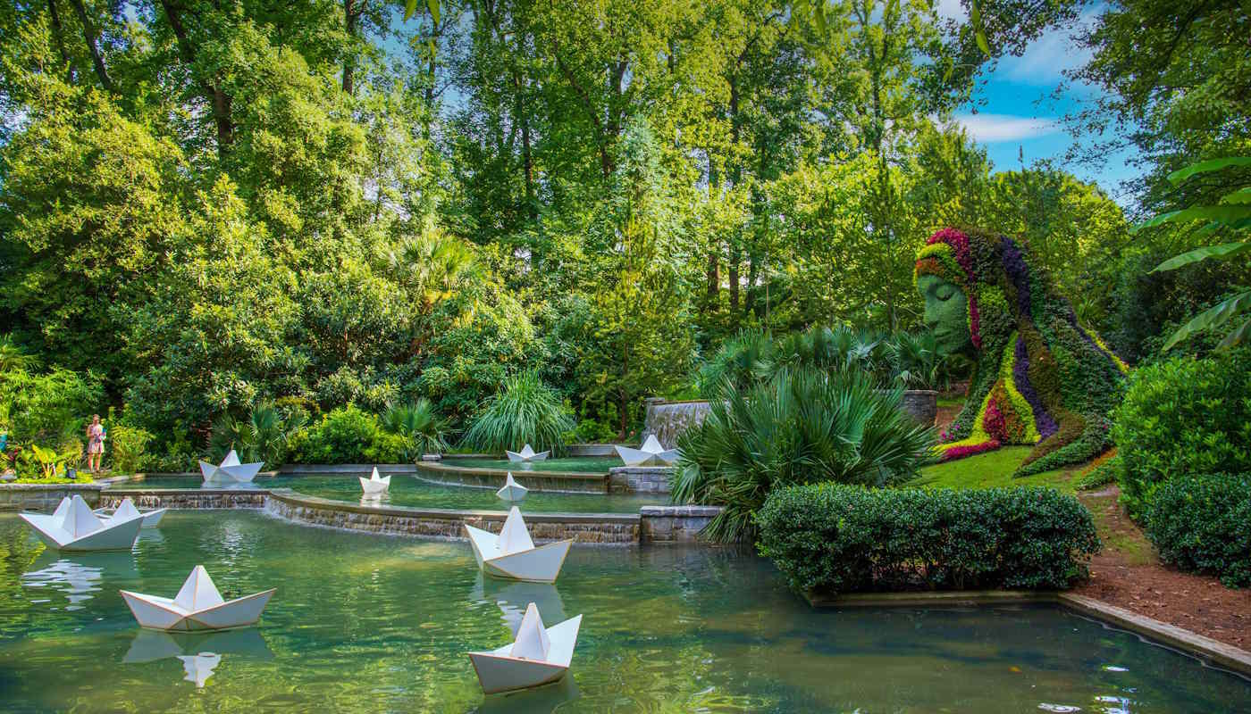 Botanischer Garten in Atlanta, USA Im Botanischen Garten von Atlanta erstreckt sich eine üppig grüne Gartenlandschaft mit ruhigen Wasserbecken, auf denen mehrere weiße Papierboote treiben. Ein besonderer Blickfang ist die große, aus bunten Pflanzen gestaltete Skulptur einer sitzenden Figur, die dem Garten eine märchenhafte und kunstvolle Atmosphäre verleiht.