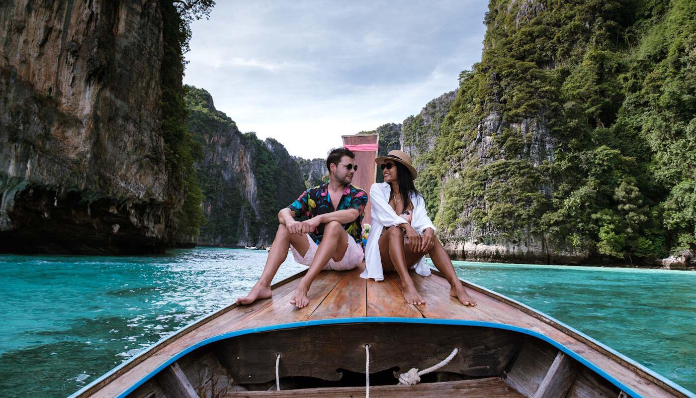 Insel Koh Phi Phi in Thailand Zwei Reisende sitzen auf dem Bug eines traditionellen Holzbootes auf der Insel Koh Phi Phi in Thailand, das durch türkisblaues Wasser zwischen steilen, üppig bewachsenen Kalksteinfelsen fährt.