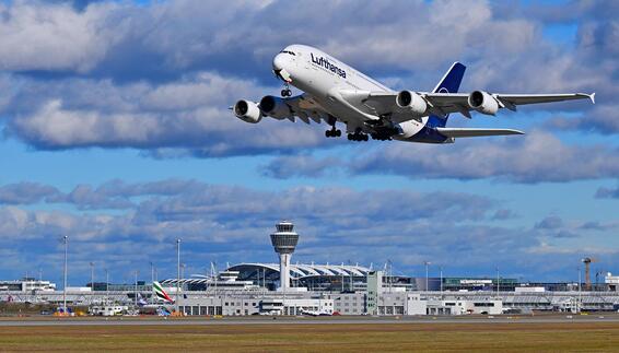 Lufthansa Airbus A380-800 beim Start über dem Flughafen München mit Terminalgebäuden und Tower im Hintergrund.