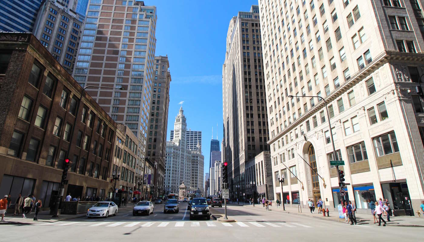 Innenstadt von Chicago, USA Eine breite Straße in der Innenstadt von Chicago wird von hohen Gebäuden aus Glas, Stein und Backstein gesäumt, die eine typische Großstadtkulisse mit markanter Skyline bilden. Auf der Fahrbahn sind mehrere Autos unterwegs, während Fußgänger die Kreuzung überqueren; im Hintergrund ragen weitere Wolkenkratzer vor einem klaren blauen Himmel auf.