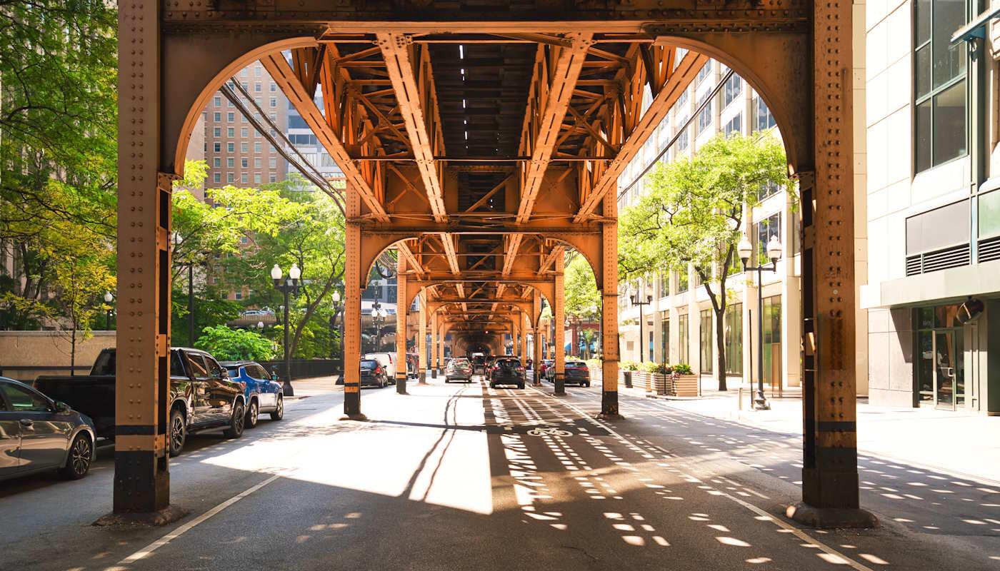 Subway Loop in Chicago, USA Das Bild zeigt die berühmte Hochbahn „L“ in der Innenstadt von Chicago, deren stählerne Trasse über einer Straße zwischen modernen Gebäuden verläuft. Sonnenlicht fällt durch die Konstruktion auf die Fahrbahn.