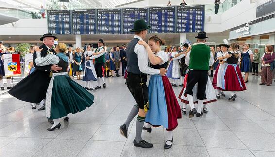 Group of people in traditional Bavarian dress dancing in an airport terminal, with a large departure board and waiting travelers in the background.