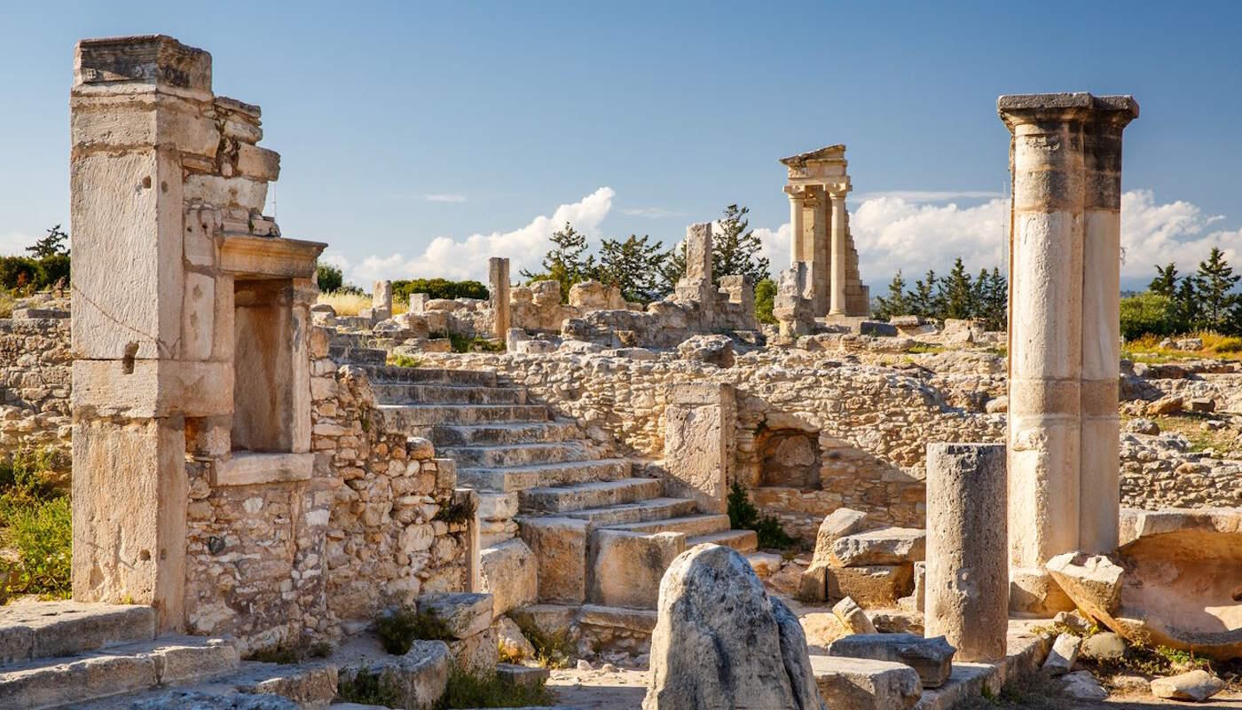 Antike Steinruinen mit hohen Säulen, Treppen und historischen Mauerresten erstrecken sich unter einem klaren blauen Himmel. Die sonnenbeschienene Ausgrabungsstätte in Kourion auf Zypern vermittelt einen Eindruck der jahrtausendealten Geschichte der Insel. Antike Ausgrabungsstätte in Kourion, Zypern